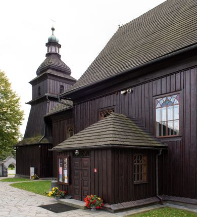 Zdjęcie nr 4: Kościół orientowany, drewniany, na podmurówce z kamienia łupanego, konstrukcji zrębowej, jednonawowy, szalowany, z wieżą konstrukcji słupowej usytuowaną od strony prezbiterium, z zakrystią w przyziemiu. Nawa prostokątna, szersza od prezbiterium, z kruchtami od zachodu, południa i północy. Prezbiterium zamknięte trójbocznie. Wieża od wschodu, na planie kwadratu, o konstrukcji ze słupów pochyłych, na zewnątrz trzy kondygnacje przedzielone obiegającymi daszkami. Dolna i górna izbica o ścianach pionowych, środkowa o ścianach pochyłych; wieża zwieńczona hełmem baniastym z latarnią. Kościół przykrywa dach siodłowy, na kruchtach trójspadowy, kryty cementówką. Pod wydatnymi okapami dachów biegnie gzyms o profilu ćwierćkoliście wklęsłym z wałkiem. Więźba dachowa nad prezbiterium jest krokwiowa, a nad nawą płatwiowa. Wewnątrz znajdują się stropy z fasetami, pod którymi umieszczony jest profilowany gzyms. Arkada tęczowa o łuku spłaszczonym. Chór muzyczny wsparty jest na dwóch filarach z wybrzuszoną balustradą. Wnętrze polichromowane, ściany w kruchtach oszalowane. Do kościoła prowadzą cztery wejścia: główne od zachodu, dwa boczne od południa i północy oraz przez zakrystię od wieży, od strony północno-zachodniej. W prezbiterium znajdują się dwa okna: po stronie południowej i północnej; a w nawie dwa po stronie południowej i północnej, natomiast w wieży cztery okna prostokątne, w latarni osiem okien zamkniętych dwoma nałęczami. Kościół otoczony jest murkiem ze stacjami drogi krzyżowej i dwiema bramkami z lat 1863 i 1864.