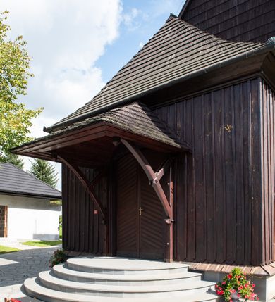 Zdjęcie nr 7: Kościół orientowany, drewniany, na podmurówce z kamienia łupanego, konstrukcji zrębowej, jednonawowy, szalowany, z wieżą konstrukcji słupowej usytuowaną od strony prezbiterium, z zakrystią w przyziemiu. Nawa prostokątna, szersza od prezbiterium, z kruchtami od zachodu, południa i północy. Prezbiterium zamknięte trójbocznie. Wieża od wschodu, na planie kwadratu, o konstrukcji ze słupów pochyłych, na zewnątrz trzy kondygnacje przedzielone obiegającymi daszkami. Dolna i górna izbica o ścianach pionowych, środkowa o ścianach pochyłych; wieża zwieńczona hełmem baniastym z latarnią. Kościół przykrywa dach siodłowy, na kruchtach trójspadowy, kryty cementówką. Pod wydatnymi okapami dachów biegnie gzyms o profilu ćwierćkoliście wklęsłym z wałkiem. Więźba dachowa nad prezbiterium jest krokwiowa, a nad nawą płatwiowa. Wewnątrz znajdują się stropy z fasetami, pod którymi umieszczony jest profilowany gzyms. Arkada tęczowa o łuku spłaszczonym. Chór muzyczny wsparty jest na dwóch filarach z wybrzuszoną balustradą. Wnętrze polichromowane, ściany w kruchtach oszalowane. Do kościoła prowadzą cztery wejścia: główne od zachodu, dwa boczne od południa i północy oraz przez zakrystię od wieży, od strony północno-zachodniej. W prezbiterium znajdują się dwa okna: po stronie południowej i północnej; a w nawie dwa po stronie południowej i północnej, natomiast w wieży cztery okna prostokątne, w latarni osiem okien zamkniętych dwoma nałęczami. Kościół otoczony jest murkiem ze stacjami drogi krzyżowej i dwiema bramkami z lat 1863 i 1864.