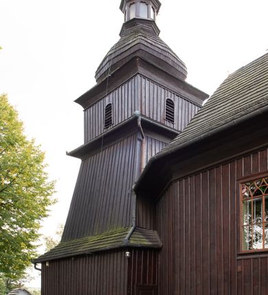 Zdjęcie nr 3: Kościół orientowany, drewniany, na podmurówce z kamienia łupanego, konstrukcji zrębowej, jednonawowy, szalowany, z wieżą konstrukcji słupowej usytuowaną od strony prezbiterium, z zakrystią w przyziemiu. Nawa prostokątna, szersza od prezbiterium, z kruchtami od zachodu, południa i północy. Prezbiterium zamknięte trójbocznie. Wieża od wschodu, na planie kwadratu, o konstrukcji ze słupów pochyłych, na zewnątrz trzy kondygnacje przedzielone obiegającymi daszkami. Dolna i górna izbica o ścianach pionowych, środkowa o ścianach pochyłych; wieża zwieńczona hełmem baniastym z latarnią. Kościół przykrywa dach siodłowy, na kruchtach trójspadowy, kryty cementówką. Pod wydatnymi okapami dachów biegnie gzyms o profilu ćwierćkoliście wklęsłym z wałkiem. Więźba dachowa nad prezbiterium jest krokwiowa, a nad nawą płatwiowa. Wewnątrz znajdują się stropy z fasetami, pod którymi umieszczony jest profilowany gzyms. Arkada tęczowa o łuku spłaszczonym. Chór muzyczny wsparty jest na dwóch filarach z wybrzuszoną balustradą. Wnętrze polichromowane, ściany w kruchtach oszalowane. Do kościoła prowadzą cztery wejścia: główne od zachodu, dwa boczne od południa i północy oraz przez zakrystię od wieży, od strony północno-zachodniej. W prezbiterium znajdują się dwa okna: po stronie południowej i północnej; a w nawie dwa po stronie południowej i północnej, natomiast w wieży cztery okna prostokątne, w latarni osiem okien zamkniętych dwoma nałęczami. Kościół otoczony jest murkiem ze stacjami drogi krzyżowej i dwiema bramkami z lat 1863 i 1864.