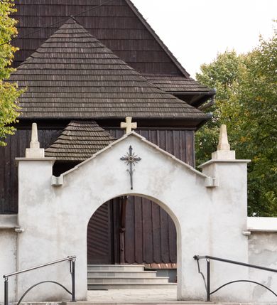 Zdjęcie nr 15: Kościół orientowany, drewniany, na podmurówce z kamienia łupanego, konstrukcji zrębowej, jednonawowy, szalowany, z wieżą konstrukcji słupowej usytuowaną od strony prezbiterium, z zakrystią w przyziemiu. Nawa prostokątna, szersza od prezbiterium, z kruchtami od zachodu, południa i północy. Prezbiterium zamknięte trójbocznie. Wieża od wschodu, na planie kwadratu, o konstrukcji ze słupów pochyłych, na zewnątrz trzy kondygnacje przedzielone obiegającymi daszkami. Dolna i górna izbica o ścianach pionowych, środkowa o ścianach pochyłych; wieża zwieńczona hełmem baniastym z latarnią. Kościół przykrywa dach siodłowy, na kruchtach trójspadowy, kryty cementówką. Pod wydatnymi okapami dachów biegnie gzyms o profilu ćwierćkoliście wklęsłym z wałkiem. Więźba dachowa nad prezbiterium jest krokwiowa, a nad nawą płatwiowa. Wewnątrz znajdują się stropy z fasetami, pod którymi umieszczony jest profilowany gzyms. Arkada tęczowa o łuku spłaszczonym. Chór muzyczny wsparty jest na dwóch filarach z wybrzuszoną balustradą. Wnętrze polichromowane, ściany w kruchtach oszalowane. Do kościoła prowadzą cztery wejścia: główne od zachodu, dwa boczne od południa i północy oraz przez zakrystię od wieży, od strony północno-zachodniej. W prezbiterium znajdują się dwa okna: po stronie południowej i północnej; a w nawie dwa po stronie południowej i północnej, natomiast w wieży cztery okna prostokątne, w latarni osiem okien zamkniętych dwoma nałęczami. Kościół otoczony jest murkiem ze stacjami drogi krzyżowej i dwiema bramkami z lat 1863 i 1864.