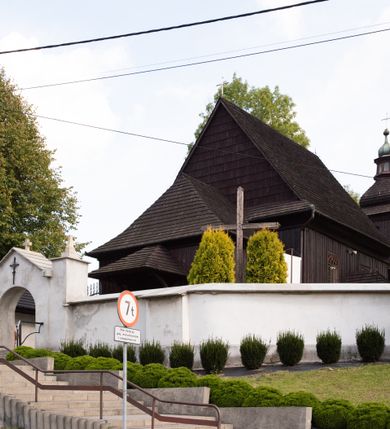 Zdjęcie nr 16: Kościół orientowany, drewniany, na podmurówce z kamienia łupanego, konstrukcji zrębowej, jednonawowy, szalowany, z wieżą konstrukcji słupowej usytuowaną od strony prezbiterium, z zakrystią w przyziemiu. Nawa prostokątna, szersza od prezbiterium, z kruchtami od zachodu, południa i północy. Prezbiterium zamknięte trójbocznie. Wieża od wschodu, na planie kwadratu, o konstrukcji ze słupów pochyłych, na zewnątrz trzy kondygnacje przedzielone obiegającymi daszkami. Dolna i górna izbica o ścianach pionowych, środkowa o ścianach pochyłych; wieża zwieńczona hełmem baniastym z latarnią. Kościół przykrywa dach siodłowy, na kruchtach trójspadowy, kryty cementówką. Pod wydatnymi okapami dachów biegnie gzyms o profilu ćwierćkoliście wklęsłym z wałkiem. Więźba dachowa nad prezbiterium jest krokwiowa, a nad nawą płatwiowa. Wewnątrz znajdują się stropy z fasetami, pod którymi umieszczony jest profilowany gzyms. Arkada tęczowa o łuku spłaszczonym. Chór muzyczny wsparty jest na dwóch filarach z wybrzuszoną balustradą. Wnętrze polichromowane, ściany w kruchtach oszalowane. Do kościoła prowadzą cztery wejścia: główne od zachodu, dwa boczne od południa i północy oraz przez zakrystię od wieży, od strony północno-zachodniej. W prezbiterium znajdują się dwa okna: po stronie południowej i północnej; a w nawie dwa po stronie południowej i północnej, natomiast w wieży cztery okna prostokątne, w latarni osiem okien zamkniętych dwoma nałęczami. Kościół otoczony jest murkiem ze stacjami drogi krzyżowej i dwiema bramkami z lat 1863 i 1864.