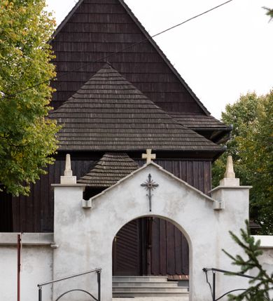 Zdjęcie nr 14: Kościół orientowany, drewniany, na podmurówce z kamienia łupanego, konstrukcji zrębowej, jednonawowy, szalowany, z wieżą konstrukcji słupowej usytuowaną od strony prezbiterium, z zakrystią w przyziemiu. Nawa prostokątna, szersza od prezbiterium, z kruchtami od zachodu, południa i północy. Prezbiterium zamknięte trójbocznie. Wieża od wschodu, na planie kwadratu, o konstrukcji ze słupów pochyłych, na zewnątrz trzy kondygnacje przedzielone obiegającymi daszkami. Dolna i górna izbica o ścianach pionowych, środkowa o ścianach pochyłych; wieża zwieńczona hełmem baniastym z latarnią. Kościół przykrywa dach siodłowy, na kruchtach trójspadowy, kryty cementówką. Pod wydatnymi okapami dachów biegnie gzyms o profilu ćwierćkoliście wklęsłym z wałkiem. Więźba dachowa nad prezbiterium jest krokwiowa, a nad nawą płatwiowa. Wewnątrz znajdują się stropy z fasetami, pod którymi umieszczony jest profilowany gzyms. Arkada tęczowa o łuku spłaszczonym. Chór muzyczny wsparty jest na dwóch filarach z wybrzuszoną balustradą. Wnętrze polichromowane, ściany w kruchtach oszalowane. Do kościoła prowadzą cztery wejścia: główne od zachodu, dwa boczne od południa i północy oraz przez zakrystię od wieży, od strony północno-zachodniej. W prezbiterium znajdują się dwa okna: po stronie południowej i północnej; a w nawie dwa po stronie południowej i północnej, natomiast w wieży cztery okna prostokątne, w latarni osiem okien zamkniętych dwoma nałęczami. Kościół otoczony jest murkiem ze stacjami drogi krzyżowej i dwiema bramkami z lat 1863 i 1864.