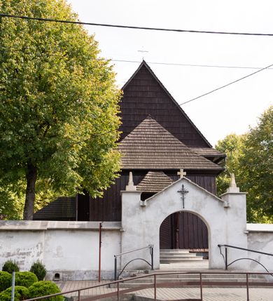 Zdjęcie nr 12: Kościół orientowany, drewniany, na podmurówce z kamienia łupanego, konstrukcji zrębowej, jednonawowy, szalowany, z wieżą konstrukcji słupowej usytuowaną od strony prezbiterium, z zakrystią w przyziemiu. Nawa prostokątna, szersza od prezbiterium, z kruchtami od zachodu, południa i północy. Prezbiterium zamknięte trójbocznie. Wieża od wschodu, na planie kwadratu, o konstrukcji ze słupów pochyłych, na zewnątrz trzy kondygnacje przedzielone obiegającymi daszkami. Dolna i górna izbica o ścianach pionowych, środkowa o ścianach pochyłych; wieża zwieńczona hełmem baniastym z latarnią. Kościół przykrywa dach siodłowy, na kruchtach trójspadowy, kryty cementówką. Pod wydatnymi okapami dachów biegnie gzyms o profilu ćwierćkoliście wklęsłym z wałkiem. Więźba dachowa nad prezbiterium jest krokwiowa, a nad nawą płatwiowa. Wewnątrz znajdują się stropy z fasetami, pod którymi umieszczony jest profilowany gzyms. Arkada tęczowa o łuku spłaszczonym. Chór muzyczny wsparty jest na dwóch filarach z wybrzuszoną balustradą. Wnętrze polichromowane, ściany w kruchtach oszalowane. Do kościoła prowadzą cztery wejścia: główne od zachodu, dwa boczne od południa i północy oraz przez zakrystię od wieży, od strony północno-zachodniej. W prezbiterium znajdują się dwa okna: po stronie południowej i północnej; a w nawie dwa po stronie południowej i północnej, natomiast w wieży cztery okna prostokątne, w latarni osiem okien zamkniętych dwoma nałęczami. Kościół otoczony jest murkiem ze stacjami drogi krzyżowej i dwiema bramkami z lat 1863 i 1864.