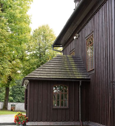 Zdjęcie nr 8: Kościół orientowany, drewniany, na podmurówce z kamienia łupanego, konstrukcji zrębowej, jednonawowy, szalowany, z wieżą konstrukcji słupowej usytuowaną od strony prezbiterium, z zakrystią w przyziemiu. Nawa prostokątna, szersza od prezbiterium, z kruchtami od zachodu, południa i północy. Prezbiterium zamknięte trójbocznie. Wieża od wschodu, na planie kwadratu, o konstrukcji ze słupów pochyłych, na zewnątrz trzy kondygnacje przedzielone obiegającymi daszkami. Dolna i górna izbica o ścianach pionowych, środkowa o ścianach pochyłych; wieża zwieńczona hełmem baniastym z latarnią. Kościół przykrywa dach siodłowy, na kruchtach trójspadowy, kryty cementówką. Pod wydatnymi okapami dachów biegnie gzyms o profilu ćwierćkoliście wklęsłym z wałkiem. Więźba dachowa nad prezbiterium jest krokwiowa, a nad nawą płatwiowa. Wewnątrz znajdują się stropy z fasetami, pod którymi umieszczony jest profilowany gzyms. Arkada tęczowa o łuku spłaszczonym. Chór muzyczny wsparty jest na dwóch filarach z wybrzuszoną balustradą. Wnętrze polichromowane, ściany w kruchtach oszalowane. Do kościoła prowadzą cztery wejścia: główne od zachodu, dwa boczne od południa i północy oraz przez zakrystię od wieży, od strony północno-zachodniej. W prezbiterium znajdują się dwa okna: po stronie południowej i północnej; a w nawie dwa po stronie południowej i północnej, natomiast w wieży cztery okna prostokątne, w latarni osiem okien zamkniętych dwoma nałęczami. Kościół otoczony jest murkiem ze stacjami drogi krzyżowej i dwiema bramkami z lat 1863 i 1864.