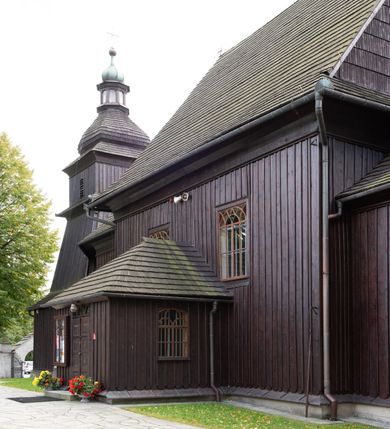 Zdjęcie nr 9: Kościół orientowany, drewniany, na podmurówce z kamienia łupanego, konstrukcji zrębowej, jednonawowy, szalowany, z wieżą konstrukcji słupowej usytuowaną od strony prezbiterium, z zakrystią w przyziemiu. Nawa prostokątna, szersza od prezbiterium, z kruchtami od zachodu, południa i północy. Prezbiterium zamknięte trójbocznie. Wieża od wschodu, na planie kwadratu, o konstrukcji ze słupów pochyłych, na zewnątrz trzy kondygnacje przedzielone obiegającymi daszkami. Dolna i górna izbica o ścianach pionowych, środkowa o ścianach pochyłych; wieża zwieńczona hełmem baniastym z latarnią. Kościół przykrywa dach siodłowy, na kruchtach trójspadowy, kryty cementówką. Pod wydatnymi okapami dachów biegnie gzyms o profilu ćwierćkoliście wklęsłym z wałkiem. Więźba dachowa nad prezbiterium jest krokwiowa, a nad nawą płatwiowa. Wewnątrz znajdują się stropy z fasetami, pod którymi umieszczony jest profilowany gzyms. Arkada tęczowa o łuku spłaszczonym. Chór muzyczny wsparty jest na dwóch filarach z wybrzuszoną balustradą. Wnętrze polichromowane, ściany w kruchtach oszalowane. Do kościoła prowadzą cztery wejścia: główne od zachodu, dwa boczne od południa i północy oraz przez zakrystię od wieży, od strony północno-zachodniej. W prezbiterium znajdują się dwa okna: po stronie południowej i północnej; a w nawie dwa po stronie południowej i północnej, natomiast w wieży cztery okna prostokątne, w latarni osiem okien zamkniętych dwoma nałęczami. Kościół otoczony jest murkiem ze stacjami drogi krzyżowej i dwiema bramkami z lat 1863 i 1864.