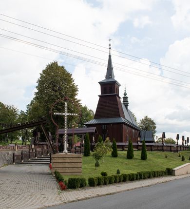 Zdjęcie nr 10: Kościół Przemienienia Pańskiego w Radoczy usytuowany jest w centrum wsi na zachód od głównej drogi prowadzącej z Wadowic do Zatora. Teren przykościelny jest usytuowany na niewielkim wzniesieniu, otoczony drewnianym płotem. Główne wejście na plac przykościelny prowadzi przez bramę od strony zachodniej.

Kościół jest orientowany, drewniany o konstrukcji zrębowej z wieżą na słup, jednonawowy z krótszym prezbiterium zamkniętym trójbocznie. Przy prezbiterium od wschodu nowsza zakrystia murowana, od północy kaplica i składzik, przy nawie od południa kruchta. Wnętrze nakryte pułapem, w nawie z zaskrzynieniami. Ściana arkady tęczowej o wykroju prostokątnym ze ściętymi narożami. Do kościoła prowadzi główne wejście w wieży od strony północnej. Wnętrze świątyni oświetlają prostokątne okna od strony południowej. Dach o jednej kalenicy, kryty blachą, na środku drewniana sygnaturka, nakryta kopułką z latarnią.  Od zachodu wieża w dolnej kondygnacji na planie prostokąta, ujęta po bokach daszkami; druga kondygnacja o ścianach pochyłych, pokryta gontem; trzecia kondygnacja prześwietlona prostokątnymi otworami okiennymi; wieża zakończona iglicą. Ściany na zewnątrz pokryte szalunkiem pionowym.
