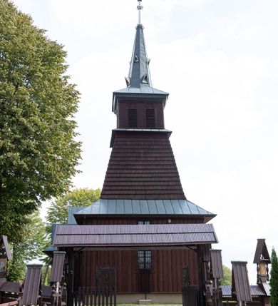 Zdjęcie nr 18: Kościół Przemienienia Pańskiego w Radoczy usytuowany jest w centrum wsi na zachód od głównej drogi prowadzącej z Wadowic do Zatora. Teren przykościelny jest usytuowany na niewielkim wzniesieniu, otoczony drewnianym płotem. Główne wejście na plac przykościelny prowadzi przez bramę od strony zachodniej.

Kościół jest orientowany, drewniany o konstrukcji zrębowej z wieżą na słup, jednonawowy z krótszym prezbiterium zamkniętym trójbocznie. Przy prezbiterium od wschodu nowsza zakrystia murowana, od północy kaplica i składzik, przy nawie od południa kruchta. Wnętrze nakryte pułapem, w nawie z zaskrzynieniami. Ściana arkady tęczowej o wykroju prostokątnym ze ściętymi narożami. Do kościoła prowadzi główne wejście w wieży od strony północnej. Wnętrze świątyni oświetlają prostokątne okna od strony południowej. Dach o jednej kalenicy, kryty blachą, na środku drewniana sygnaturka, nakryta kopułką z latarnią.  Od zachodu wieża w dolnej kondygnacji na planie prostokąta, ujęta po bokach daszkami; druga kondygnacja o ścianach pochyłych, pokryta gontem; trzecia kondygnacja prześwietlona prostokątnymi otworami okiennymi; wieża zakończona iglicą. Ściany na zewnątrz pokryte szalunkiem pionowym.
