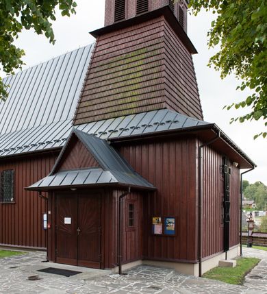 Zdjęcie nr 28: Kościół Przemienienia Pańskiego w Radoczy usytuowany jest w centrum wsi na zachód od głównej drogi prowadzącej z Wadowic do Zatora. Teren przykościelny jest usytuowany na niewielkim wzniesieniu, otoczony drewnianym płotem. Główne wejście na plac przykościelny prowadzi przez bramę od strony zachodniej.

Kościół jest orientowany, drewniany o konstrukcji zrębowej z wieżą na słup, jednonawowy z krótszym prezbiterium zamkniętym trójbocznie. Przy prezbiterium od wschodu nowsza zakrystia murowana, od północy kaplica i składzik, przy nawie od południa kruchta. Wnętrze nakryte pułapem, w nawie z zaskrzynieniami. Ściana arkady tęczowej o wykroju prostokątnym ze ściętymi narożami. Do kościoła prowadzi główne wejście w wieży od strony północnej. Wnętrze świątyni oświetlają prostokątne okna od strony południowej. Dach o jednej kalenicy, kryty blachą, na środku drewniana sygnaturka, nakryta kopułką z latarnią.  Od zachodu wieża w dolnej kondygnacji na planie prostokąta, ujęta po bokach daszkami; druga kondygnacja o ścianach pochyłych, pokryta gontem; trzecia kondygnacja prześwietlona prostokątnymi otworami okiennymi; wieża zakończona iglicą. Ściany na zewnątrz pokryte szalunkiem pionowym.
