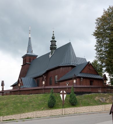 Zdjęcie nr 51: Kościół Przemienienia Pańskiego w Radoczy usytuowany jest w centrum wsi na zachód od głównej drogi prowadzącej z Wadowic do Zatora. Teren przykościelny jest usytuowany na niewielkim wzniesieniu, otoczony drewnianym płotem. Główne wejście na plac przykościelny prowadzi przez bramę od strony zachodniej.

Kościół jest orientowany, drewniany o konstrukcji zrębowej z wieżą na słup, jednonawowy z krótszym prezbiterium zamkniętym trójbocznie. Przy prezbiterium od wschodu nowsza zakrystia murowana, od północy kaplica i składzik, przy nawie od południa kruchta. Wnętrze nakryte pułapem, w nawie z zaskrzynieniami. Ściana arkady tęczowej o wykroju prostokątnym ze ściętymi narożami. Do kościoła prowadzi główne wejście w wieży od strony północnej. Wnętrze świątyni oświetlają prostokątne okna od strony południowej. Dach o jednej kalenicy, kryty blachą, na środku drewniana sygnaturka, nakryta kopułką z latarnią.  Od zachodu wieża w dolnej kondygnacji na planie prostokąta, ujęta po bokach daszkami; druga kondygnacja o ścianach pochyłych, pokryta gontem; trzecia kondygnacja prześwietlona prostokątnymi otworami okiennymi; wieża zakończona iglicą. Ściany na zewnątrz pokryte szalunkiem pionowym.
