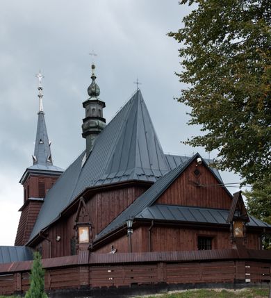 Zdjęcie nr 50: Kościół Przemienienia Pańskiego w Radoczy usytuowany jest w centrum wsi na zachód od głównej drogi prowadzącej z Wadowic do Zatora. Teren przykościelny jest usytuowany na niewielkim wzniesieniu, otoczony drewnianym płotem. Główne wejście na plac przykościelny prowadzi przez bramę od strony zachodniej.

Kościół jest orientowany, drewniany o konstrukcji zrębowej z wieżą na słup, jednonawowy z krótszym prezbiterium zamkniętym trójbocznie. Przy prezbiterium od wschodu nowsza zakrystia murowana, od północy kaplica i składzik, przy nawie od południa kruchta. Wnętrze nakryte pułapem, w nawie z zaskrzynieniami. Ściana arkady tęczowej o wykroju prostokątnym ze ściętymi narożami. Do kościoła prowadzi główne wejście w wieży od strony północnej. Wnętrze świątyni oświetlają prostokątne okna od strony południowej. Dach o jednej kalenicy, kryty blachą, na środku drewniana sygnaturka, nakryta kopułką z latarnią.  Od zachodu wieża w dolnej kondygnacji na planie prostokąta, ujęta po bokach daszkami; druga kondygnacja o ścianach pochyłych, pokryta gontem; trzecia kondygnacja prześwietlona prostokątnymi otworami okiennymi; wieża zakończona iglicą. Ściany na zewnątrz pokryte szalunkiem pionowym.
