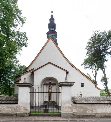 Zdjęcie nr 27: Kościół orientowany, murowany z kamienia, jednonawowy, z prostokątną, trójprzęsłową nawą oraz węższym dwuprzęsłowym prezbiterium zamkniętym prosto, z prostokątną zakrystią przy północnej ścianie. Od zachodu wieża na planie kwadratu, do północnej ściany nawy dobudowana kaplica na rzucie kwadratu o ściętych narożach, do południowej niewielka kruchta na planie kwadratu. Prezbiterium nakryte sklepieniem krzyżowo-żebrowym z dwoma zwornikami. 
Nawa sklepiona krzyżowo, przęsła wydzielone arkadami wspartymi na półfilarach. Otwór tęczowy w formie stojącego prostokąta zamkniętego łukiem półkolistym. W zachodniej części nawy parapet chóru muzycznego, z wybrzuszoną środkową częścią balustrady, wsparty na czterech kolumnach. 
Fasada wieżowa, z wejściem na osi. Wieża smukła, bez podziałów architektonicznych, zakończona profilowanym gzymsem, wyłamującym się na każdej ścianie na kształt łuku odcinkowego. W najwyższej partii ścian, z trzech stron, okna w formie stojącego prostokąta zamkniętego półkoliście. Wieża zwieńczona hełmem baniastym z latarnią, pokrytym gontem. 
Kościół opięty szkarpami dwuuskokowymi, nakrytymi daszkami gontowymi. W elewacji południowej pomiędzy szkarpami pięć wąskich okien w formie stojącego prostokąta zamkniętego łukiem półkolistym. Na wschodniej dobudowany ogrojec w formie masywnej arkady, zamkniętej dwuspadowym daszkiem z ołtarzem i krucyfiksem. Nawa i prezbiterium nakryte osobnymi dachami dwuspadowymi, gontowymi. Nad prezbiterium drewniana wieżyczka na sygnaturkę, z latarnią, zwieńczona hełmem baniastym. Kaplica przykryta dachem gontowych, trójspadowym, kruchta zaś pulpitowym. 
Kościół otoczony niskim murem kamiennym, nakrytym daszkiem gontowym. Od wschodu dwie bramy flankowane filarami z wazonami. 

