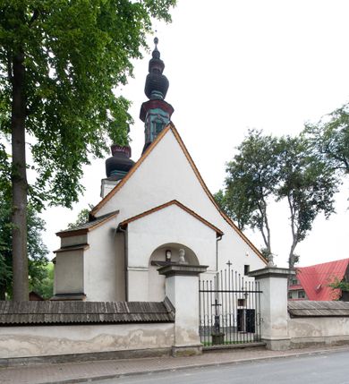 Zdjęcie nr 24: Kościół orientowany, murowany z kamienia, jednonawowy, z prostokątną, trójprzęsłową nawą oraz węższym dwuprzęsłowym prezbiterium zamkniętym prosto, z prostokątną zakrystią przy północnej ścianie. Od zachodu wieża na planie kwadratu, do północnej ściany nawy dobudowana kaplica na rzucie kwadratu o ściętych narożach, do południowej niewielka kruchta na planie kwadratu. Prezbiterium nakryte sklepieniem krzyżowo-żebrowym z dwoma zwornikami. 
Nawa sklepiona krzyżowo, przęsła wydzielone arkadami wspartymi na półfilarach. Otwór tęczowy w formie stojącego prostokąta zamkniętego łukiem półkolistym. W zachodniej części nawy parapet chóru muzycznego, z wybrzuszoną środkową częścią balustrady, wsparty na czterech kolumnach. 
Fasada wieżowa, z wejściem na osi. Wieża smukła, bez podziałów architektonicznych, zakończona profilowanym gzymsem, wyłamującym się na każdej ścianie na kształt łuku odcinkowego. W najwyższej partii ścian, z trzech stron, okna w formie stojącego prostokąta zamkniętego półkoliście. Wieża zwieńczona hełmem baniastym z latarnią, pokrytym gontem. 
Kościół opięty szkarpami dwuuskokowymi, nakrytymi daszkami gontowymi. W elewacji południowej pomiędzy szkarpami pięć wąskich okien w formie stojącego prostokąta zamkniętego łukiem półkolistym. Na wschodniej dobudowany ogrojec w formie masywnej arkady, zamkniętej dwuspadowym daszkiem z ołtarzem i krucyfiksem. Nawa i prezbiterium nakryte osobnymi dachami dwuspadowymi, gontowymi. Nad prezbiterium drewniana wieżyczka na sygnaturkę, z latarnią, zwieńczona hełmem baniastym. Kaplica przykryta dachem gontowych, trójspadowym, kruchta zaś pulpitowym. 
Kościół otoczony niskim murem kamiennym, nakrytym daszkiem gontowym. Od wschodu dwie bramy flankowane filarami z wazonami. 


