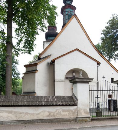 Zdjęcie nr 28: Kościół orientowany, murowany z kamienia, jednonawowy, z prostokątną, trójprzęsłową nawą oraz węższym dwuprzęsłowym prezbiterium zamkniętym prosto, z prostokątną zakrystią przy północnej ścianie. Od zachodu wieża na planie kwadratu, do północnej ściany nawy dobudowana kaplica na rzucie kwadratu o ściętych narożach, do południowej niewielka kruchta na planie kwadratu. Prezbiterium nakryte sklepieniem krzyżowo-żebrowym z dwoma zwornikami. 
Nawa sklepiona krzyżowo, przęsła wydzielone arkadami wspartymi na półfilarach. Otwór tęczowy w formie stojącego prostokąta zamkniętego łukiem półkolistym. W zachodniej części nawy parapet chóru muzycznego, z wybrzuszoną środkową częścią balustrady, wsparty na czterech kolumnach. 
Fasada wieżowa, z wejściem na osi. Wieża smukła, bez podziałów architektonicznych, zakończona profilowanym gzymsem, wyłamującym się na każdej ścianie na kształt łuku odcinkowego. W najwyższej partii ścian, z trzech stron, okna w formie stojącego prostokąta zamkniętego półkoliście. Wieża zwieńczona hełmem baniastym z latarnią, pokrytym gontem. 
Kościół opięty szkarpami dwuuskokowymi, nakrytymi daszkami gontowymi. W elewacji południowej pomiędzy szkarpami pięć wąskich okien w formie stojącego prostokąta zamkniętego łukiem półkolistym. Na wschodniej dobudowany ogrojec w formie masywnej arkady, zamkniętej dwuspadowym daszkiem z ołtarzem i krucyfiksem. Nawa i prezbiterium nakryte osobnymi dachami dwuspadowymi, gontowymi. Nad prezbiterium drewniana wieżyczka na sygnaturkę, z latarnią, zwieńczona hełmem baniastym. Kaplica przykryta dachem gontowych, trójspadowym, kruchta zaś pulpitowym. 
Kościół otoczony niskim murem kamiennym, nakrytym daszkiem gontowym. Od wschodu dwie bramy flankowane filarami z wazonami. 

