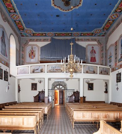 Zdjęcie nr 12: Kościół orientowany, jednonawowy, z krótkim zakończonym wielobocznie prezbiterium, przy którym od północy zakrystia z nowszym przedsionkiem; przy nawie od północy prostokątna kaplica Różańcowa (1712 r.), od zachodu wieża na rzucie kwadratu, od południa drewniana kruchta. Wewnątrz stropy. Arkada tęczy szeroka półkolista. Zewnątrz mury opięte przyporami, tynkowane. Dachy dwuspadowe i pulpitowe. Na kalenicy dachu nawy wieżyczka na sygnaturkę z datą 1878. Okna prostokątne zamknięte półkoliście. Dwa portale kamienne prostokątne. Wieża dołem gotycka, nadbudowana w stylu pseudoromańskim; trójkondygnacyjna, dołem z narożnikami artykułowanymi boniowaniem, narożniki górnej kondygnacji ujęte lizenami połączonymi fryzem arkadkowym. Elewacje wieży zwieńczone trójkątnymi szczytami i ostrosłupowym hełmem krytym blachą.