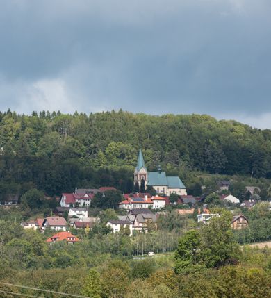 Zdjęcie nr 20: Kościół orientowany, jednonawowy, z krótkim zakończonym wielobocznie prezbiterium, przy którym od północy zakrystia z nowszym przedsionkiem; przy nawie od północy prostokątna kaplica Różańcowa (1712 r.), od zachodu wieża na rzucie kwadratu, od południa drewniana kruchta. Wewnątrz stropy. Arkada tęczy szeroka półkolista. Zewnątrz mury opięte przyporami, tynkowane. Dachy dwuspadowe i pulpitowe. Na kalenicy dachu nawy wieżyczka na sygnaturkę z datą 1878. Okna prostokątne zamknięte półkoliście. Dwa portale kamienne prostokątne. Wieża dołem gotycka, nadbudowana w stylu pseudoromańskim; trójkondygnacyjna, dołem z narożnikami artykułowanymi boniowaniem, narożniki górnej kondygnacji ujęte lizenami połączonymi fryzem arkadkowym. Elewacje wieży zwieńczone trójkątnymi szczytami i ostrosłupowym hełmem krytym blachą.