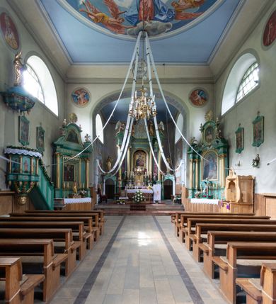 Zdjęcie nr 19: Kościół murowany, składa się z nawy i zwróconego na południe prezbiterium, do którego przylega zakrystia, nad którą znajduje się skarbiec; od północy wieża. Prezbiterium jest węższe od nawy, o zaokrąglonych narożach, oddzielone od prostokątnej nawy łukiem tęczowym. Ściany kościoła gładkie, przeprute półkolistymi oknami – dwoma w prezbiterium, sześcioma w nawie. Od zachodu chór wsparty na dwóch kolumnach, z balustradą wyłamaną na środku, dekorowaną u spodu lambrekinem. Ściany kościoła gładkie, w dolnej partii osłonięte drewnianą boazerią, z wnęką na chrzcielnicę po lewej i drugą na drzwi z prawej strony. Wnętrze przekrywają stropy – zwierciadlany w prezbiterium i płaski  z fasetami w nawie. Na nich oraz w górnej partii ścian polichromia. Nad prezbiterium wizerunek zmartwychwstałego Chrystusa, nad nawą zakomponowana w kole scena adoracji Najświętszej Marii Panny przez apostołów, poniżej, na ścianach medaliony z postaciami ważnych osobistości, świętych i aniołów. Główne drzwi od północy, boczne od zachodu. 
Kościół posadowiony na cokole. Elewacja frontowa ze zdwojonym ryzalitem, którego środek odpowiada szerokości wieży; podziały poziome przeprowadzone przy pomocy gzymsów. Na osi wejście i  okno, oba w boniowanym obramieniu. Okno ujmuje polichromowana scena Zwiastowania. Ryzalit wieńczy trójkątny fronton. Cofnięty ryzalit w dolnej partii boniowany, wyżej gładki, zwieńczony ćwierćkolistymi fragmentami przerwanego przyczółka. Boki elewacji frontowej gładkie, zwieńczone frontonem przerwanym na masyw czworobocznej wieży przeprutej z każdej strony oknem w kształcie stojącego prostokąta zamkniętego łukiem pełnym. Elewacje boczne gładkie, przeprute półkolistymi oknami. Od wschodu poniżej okien szare płyciny, od południa, w kapliczkowej oprawie grupa Ukrzyżowania. Dach nad nawą dwuspadowy, nad prezbiterium trójspadowy, dach wieńczy baniasty hełm z latarnią zakończony krzyżem, na kalenicy barokowa wieżyczka na sygnaturkę. 
