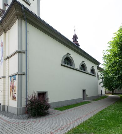 Zdjęcie nr 9: Kościół murowany, składa się z nawy i zwróconego na południe prezbiterium, do którego przylega zakrystia, nad którą znajduje się skarbiec; od północy wieża. Prezbiterium jest węższe od nawy, o zaokrąglonych narożach, oddzielone od prostokątnej nawy łukiem tęczowym. Ściany kościoła gładkie, przeprute półkolistymi oknami – dwoma w prezbiterium, sześcioma w nawie. Od zachodu chór wsparty na dwóch kolumnach, z balustradą wyłamaną na środku, dekorowaną u spodu lambrekinem. Ściany kościoła gładkie, w dolnej partii osłonięte drewnianą boazerią, z wnęką na chrzcielnicę po lewej i drugą na drzwi z prawej strony. Wnętrze przekrywają stropy – zwierciadlany w prezbiterium i płaski  z fasetami w nawie. Na nich oraz w górnej partii ścian polichromia. Nad prezbiterium wizerunek zmartwychwstałego Chrystusa, nad nawą zakomponowana w kole scena adoracji Najświętszej Marii Panny przez apostołów, poniżej, na ścianach medaliony z postaciami ważnych osobistości, świętych i aniołów. Główne drzwi od północy, boczne od zachodu. 
Kościół posadowiony na cokole. Elewacja frontowa ze zdwojonym ryzalitem, którego środek odpowiada szerokości wieży; podziały poziome przeprowadzone przy pomocy gzymsów. Na osi wejście i  okno, oba w boniowanym obramieniu. Okno ujmuje polichromowana scena Zwiastowania. Ryzalit wieńczy trójkątny fronton. Cofnięty ryzalit w dolnej partii boniowany, wyżej gładki, zwieńczony ćwierćkolistymi fragmentami przerwanego przyczółka. Boki elewacji frontowej gładkie, zwieńczone frontonem przerwanym na masyw czworobocznej wieży przeprutej z każdej strony oknem w kształcie stojącego prostokąta zamkniętego łukiem pełnym. Elewacje boczne gładkie, przeprute półkolistymi oknami. Od wschodu poniżej okien szare płyciny, od południa, w kapliczkowej oprawie grupa Ukrzyżowania. Dach nad nawą dwuspadowy, nad prezbiterium trójspadowy, dach wieńczy baniasty hełm z latarnią zakończony krzyżem, na kalenicy barokowa wieżyczka na sygnaturkę. 
