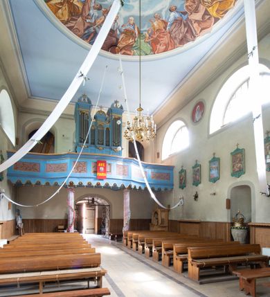 Zdjęcie nr 26: Kościół murowany, składa się z nawy i zwróconego na południe prezbiterium, do którego przylega zakrystia, nad którą znajduje się skarbiec; od północy wieża. Prezbiterium jest węższe od nawy, o zaokrąglonych narożach, oddzielone od prostokątnej nawy łukiem tęczowym. Ściany kościoła gładkie, przeprute półkolistymi oknami – dwoma w prezbiterium, sześcioma w nawie. Od zachodu chór wsparty na dwóch kolumnach, z balustradą wyłamaną na środku, dekorowaną u spodu lambrekinem. Ściany kościoła gładkie, w dolnej partii osłonięte drewnianą boazerią, z wnęką na chrzcielnicę po lewej i drugą na drzwi z prawej strony. Wnętrze przekrywają stropy – zwierciadlany w prezbiterium i płaski  z fasetami w nawie. Na nich oraz w górnej partii ścian polichromia. Nad prezbiterium wizerunek zmartwychwstałego Chrystusa, nad nawą zakomponowana w kole scena adoracji Najświętszej Marii Panny przez apostołów, poniżej, na ścianach medaliony z postaciami ważnych osobistości, świętych i aniołów. Główne drzwi od północy, boczne od zachodu. 
Kościół posadowiony na cokole. Elewacja frontowa ze zdwojonym ryzalitem, którego środek odpowiada szerokości wieży; podziały poziome przeprowadzone przy pomocy gzymsów. Na osi wejście i  okno, oba w boniowanym obramieniu. Okno ujmuje polichromowana scena Zwiastowania. Ryzalit wieńczy trójkątny fronton. Cofnięty ryzalit w dolnej partii boniowany, wyżej gładki, zwieńczony ćwierćkolistymi fragmentami przerwanego przyczółka. Boki elewacji frontowej gładkie, zwieńczone frontonem przerwanym na masyw czworobocznej wieży przeprutej z każdej strony oknem w kształcie stojącego prostokąta zamkniętego łukiem pełnym. Elewacje boczne gładkie, przeprute półkolistymi oknami. Od wschodu poniżej okien szare płyciny, od południa, w kapliczkowej oprawie grupa Ukrzyżowania. Dach nad nawą dwuspadowy, nad prezbiterium trójspadowy, dach wieńczy baniasty hełm z latarnią zakończony krzyżem, na kalenicy barokowa wieżyczka na sygnaturkę. 
