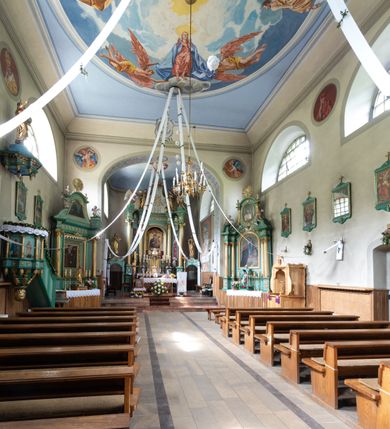 Zdjęcie nr 21: Kościół murowany, składa się z nawy i zwróconego na południe prezbiterium, do którego przylega zakrystia, nad którą znajduje się skarbiec; od północy wieża. Prezbiterium jest węższe od nawy, o zaokrąglonych narożach, oddzielone od prostokątnej nawy łukiem tęczowym. Ściany kościoła gładkie, przeprute półkolistymi oknami – dwoma w prezbiterium, sześcioma w nawie. Od zachodu chór wsparty na dwóch kolumnach, z balustradą wyłamaną na środku, dekorowaną u spodu lambrekinem. Ściany kościoła gładkie, w dolnej partii osłonięte drewnianą boazerią, z wnęką na chrzcielnicę po lewej i drugą na drzwi z prawej strony. Wnętrze przekrywają stropy – zwierciadlany w prezbiterium i płaski  z fasetami w nawie. Na nich oraz w górnej partii ścian polichromia. Nad prezbiterium wizerunek zmartwychwstałego Chrystusa, nad nawą zakomponowana w kole scena adoracji Najświętszej Marii Panny przez apostołów, poniżej, na ścianach medaliony z postaciami ważnych osobistości, świętych i aniołów. Główne drzwi od północy, boczne od zachodu. 
Kościół posadowiony na cokole. Elewacja frontowa ze zdwojonym ryzalitem, którego środek odpowiada szerokości wieży; podziały poziome przeprowadzone przy pomocy gzymsów. Na osi wejście i  okno, oba w boniowanym obramieniu. Okno ujmuje polichromowana scena Zwiastowania. Ryzalit wieńczy trójkątny fronton. Cofnięty ryzalit w dolnej partii boniowany, wyżej gładki, zwieńczony ćwierćkolistymi fragmentami przerwanego przyczółka. Boki elewacji frontowej gładkie, zwieńczone frontonem przerwanym na masyw czworobocznej wieży przeprutej z każdej strony oknem w kształcie stojącego prostokąta zamkniętego łukiem pełnym. Elewacje boczne gładkie, przeprute półkolistymi oknami. Od wschodu poniżej okien szare płyciny, od południa, w kapliczkowej oprawie grupa Ukrzyżowania. Dach nad nawą dwuspadowy, nad prezbiterium trójspadowy, dach wieńczy baniasty hełm z latarnią zakończony krzyżem, na kalenicy barokowa wieżyczka na sygnaturkę. 
