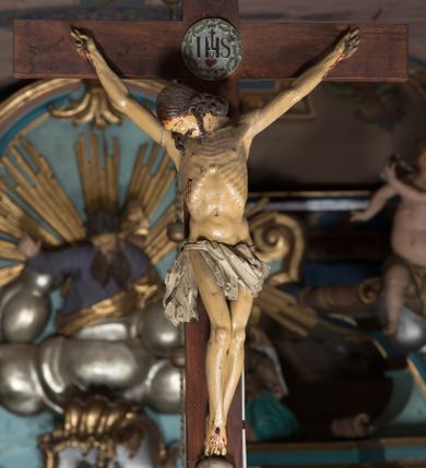 Zdjęcie nr 1: Figura umarłego Chrystusa przybita jest trzema gwoździami do prostego krzyża. Ciało w dużym zwisie, o układzie rąk bliskim literze „V”, głowa opada na prawy bark, nogi delikatnie ugięte w kolanach, stopy skrzyżowane w układzie prawa na lewą. Twarz szczupła, okolona zarostem oraz silnie kręconymi włosami opadającymi na ramiona. Ciało Chrystusa jest bardzo szczupłe, o silnie uwypuklonych żebrach klatki piersiowej. Perizonium w postaci silnie drapowanej, białej tkaniny zawieszonej na podwójnym sznurze, odsłaniającej prawe udo, ze zwisem tkaniny przy prawym boku. Polichromia ciała naturalistyczna z zaznaczonymi śladami męki, włosy brązowe. U stóp Chrystusa czaszka. Na skrzyżowaniu belek krzyża medalion ujęty koroną cierniową z monogramem IHS i gorejącym sercem w polu. Na zwieńczeniu pionowej belki krzyża banderola z napisem „IN/RI”.
