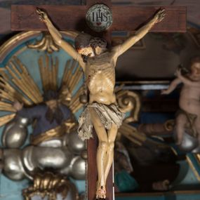 Zdjęcie nr 1: Figura umarłego Chrystusa przybita jest trzema gwoździami do prostego krzyża. Ciało w dużym zwisie, o układzie rąk bliskim literze „V”, głowa opada na prawy bark, nogi delikatnie ugięte w kolanach, stopy skrzyżowane w układzie prawa na lewą. Twarz szczupła, okolona zarostem oraz silnie kręconymi włosami opadającymi na ramiona. Ciało Chrystusa jest bardzo szczupłe, o silnie uwypuklonych żebrach klatki piersiowej. Perizonium w postaci silnie drapowanej, białej tkaniny zawieszonej na podwójnym sznurze, odsłaniającej prawe udo, ze zwisem tkaniny przy prawym boku. Polichromia ciała naturalistyczna z zaznaczonymi śladami męki, włosy brązowe. U stóp Chrystusa czaszka. Na skrzyżowaniu belek krzyża medalion ujęty koroną cierniową z monogramem IHS i gorejącym sercem w polu. Na zwieńczeniu pionowej belki krzyża banderola z napisem „IN/RI”.