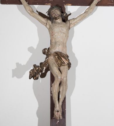 Zdjęcie nr 1: Krzyż o prostym zakończeniu ramion, ustawiony na wklęsło-wypukłej podstawie z muszlą, przytwierdzonej do drążka. Figura żywego Chrystusa przybita do krzyża trzema gwoździami. Ciało w dużym zwisie, o łukowatym układzie ramion, głowie przechylonej w prawo, esowato wygiętej sylwetce, nogi ma ugięte, a stopy skrzyżowane w układzie prawa na lewą. Chrystus ma owalną twarz, o półprzymkniętych oczach, prostym nosie i otwartych ustach, okoloną krótką brodą i silnie kręconymi, rozwianymi włosami sięgającymi ramion. Na głowie ma koronę cierniową. Ciało szczupłe, o delikatnie podkreślonej muskulaturze klatki piersiowej i z wyraźnie zaznaczonymi mięśniami ramion i nóg. Perizonium złocone, krótkie, zawieszone na podwójnym sznurze, odsłaniające prawe udo, przesłaniające partie podbrzusza i lewe biodro, o silnie rozwianym zwisie tkaniny przy prawym boku. Polichromia ciała naturalistyczna z zaznaczonymi śladami męki. Na zwieńczeniu pionowej belki krzyża srebrzona banderola z napisem „I.N.R.I.”.