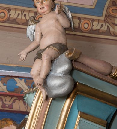 Zdjęcie nr 16: Nastawa ołtarzowa architektoniczna, jednoosiowa, jednokondygnacyjna ze zwieńczeniem, ustawiona na dwustrefowym cokole, z wyodrębnionymi dwiema parami prostopadłościanów. Ołtarz kamienny, wolnostojący, ustawiony na dwustopniowej podstawie, ujęty po bokach fryzowanymi konsolami. Mensa kamienna z marmurowym portatylem. Na przedniej ściance wyrzeźbiona w licu płycina, ujęta kanelowanymi lizenami, z monogramem IHS w polu. Na ołtarzu ustawione tabernakulum z tronem wystawienia. Tabernakulum w formie drewnianej skrzynki z obitymi blachą prostokątnymi drzwiczkami, ujętymi ramą. W ich polu medalion z przedstawieniem pelikana karmiącego młode na tle krzyża. Tron wystawienia w formie obrotowej, trójdzielnej niszy o wykroju stojącego prostokąta zamkniętego stylizowanym łukiem półokrągłym, zwieńczonej gzymsem. Nisza flankowana dekoracją w postaci liści ostrokrzewu i kampanuli, na gzymsie rocaille. Tabernakulum i tron ujęte parą niższych kolumn o kapitelach kompozytowych, na wysokich cokołach, dźwigających imposty, na których stoją wazony oraz spływami zakończonymi cokołami z fragmentami przerwanego przyczółka, na których klęczą adorujące anioły. Spływy dekorowane rocaille&#039;em z podwieszonymi kampanulami. Kondygnacja ujęta dwiema parami pilastrów i stojących przed nimi kolumn o kapitelach kompozytowych, ustawionymi delikatnie pod kątem względem lica, wewnętrzna para wysunięta do przodu. Po wewnętrznej stronie ołtarza para pilastrów, których konsole podtrzymują belkowanie, a trzony dekorowane są zwisami roślinnymi, na ich tle figury św. Joachima i św. Anny. Podpory dźwigają pełne belkowanie, wyłamane na ich osi, przerwane nad polem środkowym oraz fragmenty przerwanego, falistego przyczółka z siedzącymi puttami, a także wolutowe cokoły z wazonami na zewnętrznych krawędziach belkowania. Fryz dekorowany rozetami. W polu środkowym nisza w formie stojącego prostokąta zamkniętego łukiem półkolistym nadwieszonym, ujęta profilowaną ramą z mniejszym obrazem Matki Boskiej z Dzieciątkiem oraz przedstawieniem św. Klemensa na zasuwie. Powyżej rokokowy kartusz z monogramem Marii, na którym dwa putta trzymają koronę, z podwieszonymi zwisami kwiatowymi, ujmującymi ramę pola głównego poniżej. Zwieńczenie w formie stojącego prostokąta z wolutowymi uszakami, zamkniętego gzymsem w formie łuku odcinkowego nadwieszonego. W polu rzeźbiarskie przedstawienie półpostaci Boga Ojca pośród obłoków, na tle glorii promienistej, ujętego przez siedzące na gzymsie, adorujące aniołki. Ołtarz flankują umocowane do ścian konsole z figurami św. Piotra i św. Pawła. Struktura w całości polichromowana w kolorze jasnoniebieskim, z niebieskimi i bladoróżowymi detalami, kolumny, profile i ornamentyka złocone, detale srebrzone.  
