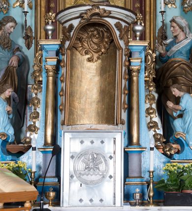Zdjęcie nr 9: Nastawa ołtarzowa architektoniczna, jednoosiowa, jednokondygnacyjna ze zwieńczeniem, ustawiona na dwustrefowym cokole, z wyodrębnionymi dwiema parami prostopadłościanów. Ołtarz kamienny, wolnostojący, ustawiony na dwustopniowej podstawie, ujęty po bokach fryzowanymi konsolami. Mensa kamienna z marmurowym portatylem. Na przedniej ściance wyrzeźbiona w licu płycina, ujęta kanelowanymi lizenami, z monogramem IHS w polu. Na ołtarzu ustawione tabernakulum z tronem wystawienia. Tabernakulum w formie drewnianej skrzynki z obitymi blachą prostokątnymi drzwiczkami, ujętymi ramą. W ich polu medalion z przedstawieniem pelikana karmiącego młode na tle krzyża. Tron wystawienia w formie obrotowej, trójdzielnej niszy o wykroju stojącego prostokąta zamkniętego stylizowanym łukiem półokrągłym, zwieńczonej gzymsem. Nisza flankowana dekoracją w postaci liści ostrokrzewu i kampanuli, na gzymsie rocaille. Tabernakulum i tron ujęte parą niższych kolumn o kapitelach kompozytowych, na wysokich cokołach, dźwigających imposty, na których stoją wazony oraz spływami zakończonymi cokołami z fragmentami przerwanego przyczółka, na których klęczą adorujące anioły. Spływy dekorowane rocaille'em z podwieszonymi kampanulami. Kondygnacja ujęta dwiema parami pilastrów i stojących przed nimi kolumn o kapitelach kompozytowych, ustawionymi delikatnie pod kątem względem lica, wewnętrzna para wysunięta do przodu. Po wewnętrznej stronie ołtarza para pilastrów, których konsole podtrzymują belkowanie, a trzony dekorowane są zwisami roślinnymi, na ich tle figury św. Joachima i św. Anny. Podpory dźwigają pełne belkowanie, wyłamane na ich osi, przerwane nad polem środkowym oraz fragmenty przerwanego, falistego przyczółka z siedzącymi puttami, a także wolutowe cokoły z wazonami na zewnętrznych krawędziach belkowania. Fryz dekorowany rozetami. W polu środkowym nisza w formie stojącego prostokąta zamkniętego łukiem półkolistym nadwieszonym, ujęta profilowaną ramą z mniejszym obrazem Matki Boskiej z Dzieciątkiem oraz przedstawieniem św. Klemensa na zasuwie. Powyżej rokokowy kartusz z monogramem Marii, na którym dwa putta trzymają koronę, z podwieszonymi zwisami kwiatowymi, ujmującymi ramę pola głównego poniżej. Zwieńczenie w formie stojącego prostokąta z wolutowymi uszakami, zamkniętego gzymsem w formie łuku odcinkowego nadwieszonego. W polu rzeźbiarskie przedstawienie półpostaci Boga Ojca pośród obłoków, na tle glorii promienistej, ujętego przez siedzące na gzymsie, adorujące aniołki. Ołtarz flankują umocowane do ścian konsole z figurami św. Piotra i św. Pawła. Struktura w całości polichromowana w kolorze jasnoniebieskim, z niebieskimi i bladoróżowymi detalami, kolumny, profile i ornamentyka złocone, detale srebrzone.  
