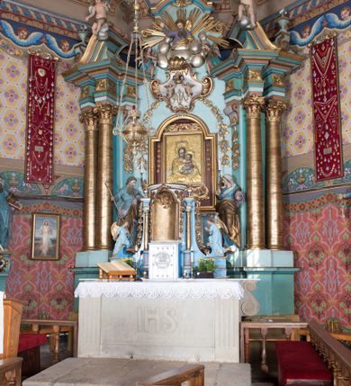 Zdjęcie nr 3: Nastawa ołtarzowa architektoniczna, jednoosiowa, jednokondygnacyjna ze zwieńczeniem, ustawiona na dwustrefowym cokole, z wyodrębnionymi dwiema parami prostopadłościanów. Ołtarz kamienny, wolnostojący, ustawiony na dwustopniowej podstawie, ujęty po bokach fryzowanymi konsolami. Mensa kamienna z marmurowym portatylem. Na przedniej ściance wyrzeźbiona w licu płycina, ujęta kanelowanymi lizenami, z monogramem IHS w polu. Na ołtarzu ustawione tabernakulum z tronem wystawienia. Tabernakulum w formie drewnianej skrzynki z obitymi blachą prostokątnymi drzwiczkami, ujętymi ramą. W ich polu medalion z przedstawieniem pelikana karmiącego młode na tle krzyża. Tron wystawienia w formie obrotowej, trójdzielnej niszy o wykroju stojącego prostokąta zamkniętego stylizowanym łukiem półokrągłym, zwieńczonej gzymsem. Nisza flankowana dekoracją w postaci liści ostrokrzewu i kampanuli, na gzymsie rocaille. Tabernakulum i tron ujęte parą niższych kolumn o kapitelach kompozytowych, na wysokich cokołach, dźwigających imposty, na których stoją wazony oraz spływami zakończonymi cokołami z fragmentami przerwanego przyczółka, na których klęczą adorujące anioły. Spływy dekorowane rocaille'em z podwieszonymi kampanulami. Kondygnacja ujęta dwiema parami pilastrów i stojących przed nimi kolumn o kapitelach kompozytowych, ustawionymi delikatnie pod kątem względem lica, wewnętrzna para wysunięta do przodu. Po wewnętrznej stronie ołtarza para pilastrów, których konsole podtrzymują belkowanie, a trzony dekorowane są zwisami roślinnymi, na ich tle figury św. Joachima i św. Anny. Podpory dźwigają pełne belkowanie, wyłamane na ich osi, przerwane nad polem środkowym oraz fragmenty przerwanego, falistego przyczółka z siedzącymi puttami, a także wolutowe cokoły z wazonami na zewnętrznych krawędziach belkowania. Fryz dekorowany rozetami. W polu środkowym nisza w formie stojącego prostokąta zamkniętego łukiem półkolistym nadwieszonym, ujęta profilowaną ramą z mniejszym obrazem Matki Boskiej z Dzieciątkiem oraz przedstawieniem św. Klemensa na zasuwie. Powyżej rokokowy kartusz z monogramem Marii, na którym dwa putta trzymają koronę, z podwieszonymi zwisami kwiatowymi, ujmującymi ramę pola głównego poniżej. Zwieńczenie w formie stojącego prostokąta z wolutowymi uszakami, zamkniętego gzymsem w formie łuku odcinkowego nadwieszonego. W polu rzeźbiarskie przedstawienie półpostaci Boga Ojca pośród obłoków, na tle glorii promienistej, ujętego przez siedzące na gzymsie, adorujące aniołki. Ołtarz flankują umocowane do ścian konsole z figurami św. Piotra i św. Pawła. Struktura w całości polichromowana w kolorze jasnoniebieskim, z niebieskimi i bladoróżowymi detalami, kolumny, profile i ornamentyka złocone, detale srebrzone.  
