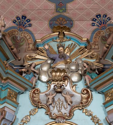 Zdjęcie nr 13: Nastawa ołtarzowa architektoniczna, jednoosiowa, jednokondygnacyjna ze zwieńczeniem, ustawiona na dwustrefowym cokole, z wyodrębnionymi dwiema parami prostopadłościanów. Ołtarz kamienny, wolnostojący, ustawiony na dwustopniowej podstawie, ujęty po bokach fryzowanymi konsolami. Mensa kamienna z marmurowym portatylem. Na przedniej ściance wyrzeźbiona w licu płycina, ujęta kanelowanymi lizenami, z monogramem IHS w polu. Na ołtarzu ustawione tabernakulum z tronem wystawienia. Tabernakulum w formie drewnianej skrzynki z obitymi blachą prostokątnymi drzwiczkami, ujętymi ramą. W ich polu medalion z przedstawieniem pelikana karmiącego młode na tle krzyża. Tron wystawienia w formie obrotowej, trójdzielnej niszy o wykroju stojącego prostokąta zamkniętego stylizowanym łukiem półokrągłym, zwieńczonej gzymsem. Nisza flankowana dekoracją w postaci liści ostrokrzewu i kampanuli, na gzymsie rocaille. Tabernakulum i tron ujęte parą niższych kolumn o kapitelach kompozytowych, na wysokich cokołach, dźwigających imposty, na których stoją wazony oraz spływami zakończonymi cokołami z fragmentami przerwanego przyczółka, na których klęczą adorujące anioły. Spływy dekorowane rocaille&#039;em z podwieszonymi kampanulami. Kondygnacja ujęta dwiema parami pilastrów i stojących przed nimi kolumn o kapitelach kompozytowych, ustawionymi delikatnie pod kątem względem lica, wewnętrzna para wysunięta do przodu. Po wewnętrznej stronie ołtarza para pilastrów, których konsole podtrzymują belkowanie, a trzony dekorowane są zwisami roślinnymi, na ich tle figury św. Joachima i św. Anny. Podpory dźwigają pełne belkowanie, wyłamane na ich osi, przerwane nad polem środkowym oraz fragmenty przerwanego, falistego przyczółka z siedzącymi puttami, a także wolutowe cokoły z wazonami na zewnętrznych krawędziach belkowania. Fryz dekorowany rozetami. W polu środkowym nisza w formie stojącego prostokąta zamkniętego łukiem półkolistym nadwieszonym, ujęta profilowaną ramą z mniejszym obrazem Matki Boskiej z Dzieciątkiem oraz przedstawieniem św. Klemensa na zasuwie. Powyżej rokokowy kartusz z monogramem Marii, na którym dwa putta trzymają koronę, z podwieszonymi zwisami kwiatowymi, ujmującymi ramę pola głównego poniżej. Zwieńczenie w formie stojącego prostokąta z wolutowymi uszakami, zamkniętego gzymsem w formie łuku odcinkowego nadwieszonego. W polu rzeźbiarskie przedstawienie półpostaci Boga Ojca pośród obłoków, na tle glorii promienistej, ujętego przez siedzące na gzymsie, adorujące aniołki. Ołtarz flankują umocowane do ścian konsole z figurami św. Piotra i św. Pawła. Struktura w całości polichromowana w kolorze jasnoniebieskim, z niebieskimi i bladoróżowymi detalami, kolumny, profile i ornamentyka złocone, detale srebrzone.  
