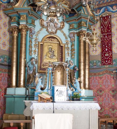 Zdjęcie nr 2: Nastawa ołtarzowa architektoniczna, jednoosiowa, jednokondygnacyjna ze zwieńczeniem, ustawiona na dwustrefowym cokole, z wyodrębnionymi dwiema parami prostopadłościanów. Ołtarz kamienny, wolnostojący, ustawiony na dwustopniowej podstawie, ujęty po bokach fryzowanymi konsolami. Mensa kamienna z marmurowym portatylem. Na przedniej ściance wyrzeźbiona w licu płycina, ujęta kanelowanymi lizenami, z monogramem IHS w polu. Na ołtarzu ustawione tabernakulum z tronem wystawienia. Tabernakulum w formie drewnianej skrzynki z obitymi blachą prostokątnymi drzwiczkami, ujętymi ramą. W ich polu medalion z przedstawieniem pelikana karmiącego młode na tle krzyża. Tron wystawienia w formie obrotowej, trójdzielnej niszy o wykroju stojącego prostokąta zamkniętego stylizowanym łukiem półokrągłym, zwieńczonej gzymsem. Nisza flankowana dekoracją w postaci liści ostrokrzewu i kampanuli, na gzymsie rocaille. Tabernakulum i tron ujęte parą niższych kolumn o kapitelach kompozytowych, na wysokich cokołach, dźwigających imposty, na których stoją wazony oraz spływami zakończonymi cokołami z fragmentami przerwanego przyczółka, na których klęczą adorujące anioły. Spływy dekorowane rocaille&#039;em z podwieszonymi kampanulami. Kondygnacja ujęta dwiema parami pilastrów i stojących przed nimi kolumn o kapitelach kompozytowych, ustawionymi delikatnie pod kątem względem lica, wewnętrzna para wysunięta do przodu. Po wewnętrznej stronie ołtarza para pilastrów, których konsole podtrzymują belkowanie, a trzony dekorowane są zwisami roślinnymi, na ich tle figury św. Joachima i św. Anny. Podpory dźwigają pełne belkowanie, wyłamane na ich osi, przerwane nad polem środkowym oraz fragmenty przerwanego, falistego przyczółka z siedzącymi puttami, a także wolutowe cokoły z wazonami na zewnętrznych krawędziach belkowania. Fryz dekorowany rozetami. W polu środkowym nisza w formie stojącego prostokąta zamkniętego łukiem półkolistym nadwieszonym, ujęta profilowaną ramą z mniejszym obrazem Matki Boskiej z Dzieciątkiem oraz przedstawieniem św. Klemensa na zasuwie. Powyżej rokokowy kartusz z monogramem Marii, na którym dwa putta trzymają koronę, z podwieszonymi zwisami kwiatowymi, ujmującymi ramę pola głównego poniżej. Zwieńczenie w formie stojącego prostokąta z wolutowymi uszakami, zamkniętego gzymsem w formie łuku odcinkowego nadwieszonego. W polu rzeźbiarskie przedstawienie półpostaci Boga Ojca pośród obłoków, na tle glorii promienistej, ujętego przez siedzące na gzymsie, adorujące aniołki. Ołtarz flankują umocowane do ścian konsole z figurami św. Piotra i św. Pawła. Struktura w całości polichromowana w kolorze jasnoniebieskim, z niebieskimi i bladoróżowymi detalami, kolumny, profile i ornamentyka złocone, detale srebrzone.  
