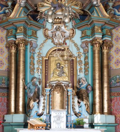 Zdjęcie nr 1: Nastawa ołtarzowa architektoniczna, jednoosiowa, jednokondygnacyjna ze zwieńczeniem, ustawiona na dwustrefowym cokole, z wyodrębnionymi dwiema parami prostopadłościanów. Ołtarz kamienny, wolnostojący, ustawiony na dwustopniowej podstawie, ujęty po bokach fryzowanymi konsolami. Mensa kamienna z marmurowym portatylem. Na przedniej ściance wyrzeźbiona w licu płycina, ujęta kanelowanymi lizenami, z monogramem IHS w polu. Na ołtarzu ustawione tabernakulum z tronem wystawienia. Tabernakulum w formie drewnianej skrzynki z obitymi blachą prostokątnymi drzwiczkami, ujętymi ramą. W ich polu medalion z przedstawieniem pelikana karmiącego młode na tle krzyża. Tron wystawienia w formie obrotowej, trójdzielnej niszy o wykroju stojącego prostokąta zamkniętego stylizowanym łukiem półokrągłym, zwieńczonej gzymsem. Nisza flankowana dekoracją w postaci liści ostrokrzewu i kampanuli, na gzymsie rocaille. Tabernakulum i tron ujęte parą niższych kolumn o kapitelach kompozytowych, na wysokich cokołach, dźwigających imposty, na których stoją wazony oraz spływami zakończonymi cokołami z fragmentami przerwanego przyczółka, na których klęczą adorujące anioły. Spływy dekorowane rocaille'em z podwieszonymi kampanulami. Kondygnacja ujęta dwiema parami pilastrów i stojących przed nimi kolumn o kapitelach kompozytowych, ustawionymi delikatnie pod kątem względem lica, wewnętrzna para wysunięta do przodu. Po wewnętrznej stronie ołtarza para pilastrów, których konsole podtrzymują belkowanie, a trzony dekorowane są zwisami roślinnymi, na ich tle figury św. Joachima i św. Anny. Podpory dźwigają pełne belkowanie, wyłamane na ich osi, przerwane nad polem środkowym oraz fragmenty przerwanego, falistego przyczółka z siedzącymi puttami, a także wolutowe cokoły z wazonami na zewnętrznych krawędziach belkowania. Fryz dekorowany rozetami. W polu środkowym nisza w formie stojącego prostokąta zamkniętego łukiem półkolistym nadwieszonym, ujęta profilowaną ramą z mniejszym obrazem Matki Boskiej z Dzieciątkiem oraz przedstawieniem św. Klemensa na zasuwie. Powyżej rokokowy kartusz z monogramem Marii, na którym dwa putta trzymają koronę, z podwieszonymi zwisami kwiatowymi, ujmującymi ramę pola głównego poniżej. Zwieńczenie w formie stojącego prostokąta z wolutowymi uszakami, zamkniętego gzymsem w formie łuku odcinkowego nadwieszonego. W polu rzeźbiarskie przedstawienie półpostaci Boga Ojca pośród obłoków, na tle glorii promienistej, ujętego przez siedzące na gzymsie, adorujące aniołki. Ołtarz flankują umocowane do ścian konsole z figurami św. Piotra i św. Pawła. Struktura w całości polichromowana w kolorze jasnoniebieskim, z niebieskimi i bladoróżowymi detalami, kolumny, profile i ornamentyka złocone, detale srebrzone.  
