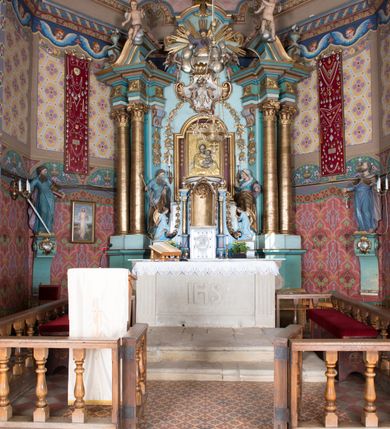 Zdjęcie nr 4: Nastawa ołtarzowa architektoniczna, jednoosiowa, jednokondygnacyjna ze zwieńczeniem, ustawiona na dwustrefowym cokole, z wyodrębnionymi dwiema parami prostopadłościanów. Ołtarz kamienny, wolnostojący, ustawiony na dwustopniowej podstawie, ujęty po bokach fryzowanymi konsolami. Mensa kamienna z marmurowym portatylem. Na przedniej ściance wyrzeźbiona w licu płycina, ujęta kanelowanymi lizenami, z monogramem IHS w polu. Na ołtarzu ustawione tabernakulum z tronem wystawienia. Tabernakulum w formie drewnianej skrzynki z obitymi blachą prostokątnymi drzwiczkami, ujętymi ramą. W ich polu medalion z przedstawieniem pelikana karmiącego młode na tle krzyża. Tron wystawienia w formie obrotowej, trójdzielnej niszy o wykroju stojącego prostokąta zamkniętego stylizowanym łukiem półokrągłym, zwieńczonej gzymsem. Nisza flankowana dekoracją w postaci liści ostrokrzewu i kampanuli, na gzymsie rocaille. Tabernakulum i tron ujęte parą niższych kolumn o kapitelach kompozytowych, na wysokich cokołach, dźwigających imposty, na których stoją wazony oraz spływami zakończonymi cokołami z fragmentami przerwanego przyczółka, na których klęczą adorujące anioły. Spływy dekorowane rocaille'em z podwieszonymi kampanulami. Kondygnacja ujęta dwiema parami pilastrów i stojących przed nimi kolumn o kapitelach kompozytowych, ustawionymi delikatnie pod kątem względem lica, wewnętrzna para wysunięta do przodu. Po wewnętrznej stronie ołtarza para pilastrów, których konsole podtrzymują belkowanie, a trzony dekorowane są zwisami roślinnymi, na ich tle figury św. Joachima i św. Anny. Podpory dźwigają pełne belkowanie, wyłamane na ich osi, przerwane nad polem środkowym oraz fragmenty przerwanego, falistego przyczółka z siedzącymi puttami, a także wolutowe cokoły z wazonami na zewnętrznych krawędziach belkowania. Fryz dekorowany rozetami. W polu środkowym nisza w formie stojącego prostokąta zamkniętego łukiem półkolistym nadwieszonym, ujęta profilowaną ramą z mniejszym obrazem Matki Boskiej z Dzieciątkiem oraz przedstawieniem św. Klemensa na zasuwie. Powyżej rokokowy kartusz z monogramem Marii, na którym dwa putta trzymają koronę, z podwieszonymi zwisami kwiatowymi, ujmującymi ramę pola głównego poniżej. Zwieńczenie w formie stojącego prostokąta z wolutowymi uszakami, zamkniętego gzymsem w formie łuku odcinkowego nadwieszonego. W polu rzeźbiarskie przedstawienie półpostaci Boga Ojca pośród obłoków, na tle glorii promienistej, ujętego przez siedzące na gzymsie, adorujące aniołki. Ołtarz flankują umocowane do ścian konsole z figurami św. Piotra i św. Pawła. Struktura w całości polichromowana w kolorze jasnoniebieskim, z niebieskimi i bladoróżowymi detalami, kolumny, profile i ornamentyka złocone, detale srebrzone.  
