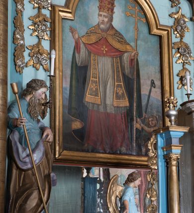 Zdjęcie nr 7: Nastawa ołtarzowa architektoniczna, jednoosiowa, jednokondygnacyjna ze zwieńczeniem, ustawiona na dwustrefowym cokole, z wyodrębnionymi dwiema parami prostopadłościanów. Ołtarz kamienny, wolnostojący, ustawiony na dwustopniowej podstawie, ujęty po bokach fryzowanymi konsolami. Mensa kamienna z marmurowym portatylem. Na przedniej ściance wyrzeźbiona w licu płycina, ujęta kanelowanymi lizenami, z monogramem IHS w polu. Na ołtarzu ustawione tabernakulum z tronem wystawienia. Tabernakulum w formie drewnianej skrzynki z obitymi blachą prostokątnymi drzwiczkami, ujętymi ramą. W ich polu medalion z przedstawieniem pelikana karmiącego młode na tle krzyża. Tron wystawienia w formie obrotowej, trójdzielnej niszy o wykroju stojącego prostokąta zamkniętego stylizowanym łukiem półokrągłym, zwieńczonej gzymsem. Nisza flankowana dekoracją w postaci liści ostrokrzewu i kampanuli, na gzymsie rocaille. Tabernakulum i tron ujęte parą niższych kolumn o kapitelach kompozytowych, na wysokich cokołach, dźwigających imposty, na których stoją wazony oraz spływami zakończonymi cokołami z fragmentami przerwanego przyczółka, na których klęczą adorujące anioły. Spływy dekorowane rocaille'em z podwieszonymi kampanulami. Kondygnacja ujęta dwiema parami pilastrów i stojących przed nimi kolumn o kapitelach kompozytowych, ustawionymi delikatnie pod kątem względem lica, wewnętrzna para wysunięta do przodu. Po wewnętrznej stronie ołtarza para pilastrów, których konsole podtrzymują belkowanie, a trzony dekorowane są zwisami roślinnymi, na ich tle figury św. Joachima i św. Anny. Podpory dźwigają pełne belkowanie, wyłamane na ich osi, przerwane nad polem środkowym oraz fragmenty przerwanego, falistego przyczółka z siedzącymi puttami, a także wolutowe cokoły z wazonami na zewnętrznych krawędziach belkowania. Fryz dekorowany rozetami. W polu środkowym nisza w formie stojącego prostokąta zamkniętego łukiem półkolistym nadwieszonym, ujęta profilowaną ramą z mniejszym obrazem Matki Boskiej z Dzieciątkiem oraz przedstawieniem św. Klemensa na zasuwie. Powyżej rokokowy kartusz z monogramem Marii, na którym dwa putta trzymają koronę, z podwieszonymi zwisami kwiatowymi, ujmującymi ramę pola głównego poniżej. Zwieńczenie w formie stojącego prostokąta z wolutowymi uszakami, zamkniętego gzymsem w formie łuku odcinkowego nadwieszonego. W polu rzeźbiarskie przedstawienie półpostaci Boga Ojca pośród obłoków, na tle glorii promienistej, ujętego przez siedzące na gzymsie, adorujące aniołki. Ołtarz flankują umocowane do ścian konsole z figurami św. Piotra i św. Pawła. Struktura w całości polichromowana w kolorze jasnoniebieskim, z niebieskimi i bladoróżowymi detalami, kolumny, profile i ornamentyka złocone, detale srebrzone.  
