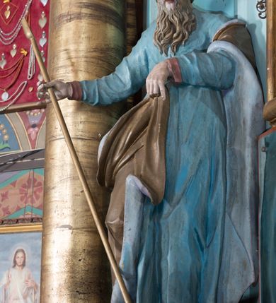 Zdjęcie nr 38: Nastawa ołtarzowa architektoniczna, jednoosiowa, jednokondygnacyjna ze zwieńczeniem, ustawiona na dwustrefowym cokole, z wyodrębnionymi dwiema parami prostopadłościanów. Ołtarz kamienny, wolnostojący, ustawiony na dwustopniowej podstawie, ujęty po bokach fryzowanymi konsolami. Mensa kamienna z marmurowym portatylem. Na przedniej ściance wyrzeźbiona w licu płycina, ujęta kanelowanymi lizenami, z monogramem IHS w polu. Na ołtarzu ustawione tabernakulum z tronem wystawienia. Tabernakulum w formie drewnianej skrzynki z obitymi blachą prostokątnymi drzwiczkami, ujętymi ramą. W ich polu medalion z przedstawieniem pelikana karmiącego młode na tle krzyża. Tron wystawienia w formie obrotowej, trójdzielnej niszy o wykroju stojącego prostokąta zamkniętego stylizowanym łukiem półokrągłym, zwieńczonej gzymsem. Nisza flankowana dekoracją w postaci liści ostrokrzewu i kampanuli, na gzymsie rocaille. Tabernakulum i tron ujęte parą niższych kolumn o kapitelach kompozytowych, na wysokich cokołach, dźwigających imposty, na których stoją wazony oraz spływami zakończonymi cokołami z fragmentami przerwanego przyczółka, na których klęczą adorujące anioły. Spływy dekorowane rocaille'em z podwieszonymi kampanulami. Kondygnacja ujęta dwiema parami pilastrów i stojących przed nimi kolumn o kapitelach kompozytowych, ustawionymi delikatnie pod kątem względem lica, wewnętrzna para wysunięta do przodu. Po wewnętrznej stronie ołtarza para pilastrów, których konsole podtrzymują belkowanie, a trzony dekorowane są zwisami roślinnymi, na ich tle figury św. Joachima i św. Anny. Podpory dźwigają pełne belkowanie, wyłamane na ich osi, przerwane nad polem środkowym oraz fragmenty przerwanego, falistego przyczółka z siedzącymi puttami, a także wolutowe cokoły z wazonami na zewnętrznych krawędziach belkowania. Fryz dekorowany rozetami. W polu środkowym nisza w formie stojącego prostokąta zamkniętego łukiem półkolistym nadwieszonym, ujęta profilowaną ramą z mniejszym obrazem Matki Boskiej z Dzieciątkiem oraz przedstawieniem św. Klemensa na zasuwie. Powyżej rokokowy kartusz z monogramem Marii, na którym dwa putta trzymają koronę, z podwieszonymi zwisami kwiatowymi, ujmującymi ramę pola głównego poniżej. Zwieńczenie w formie stojącego prostokąta z wolutowymi uszakami, zamkniętego gzymsem w formie łuku odcinkowego nadwieszonego. W polu rzeźbiarskie przedstawienie półpostaci Boga Ojca pośród obłoków, na tle glorii promienistej, ujętego przez siedzące na gzymsie, adorujące aniołki. Ołtarz flankują umocowane do ścian konsole z figurami św. Piotra i św. Pawła. Struktura w całości polichromowana w kolorze jasnoniebieskim, z niebieskimi i bladoróżowymi detalami, kolumny, profile i ornamentyka złocone, detale srebrzone.  
