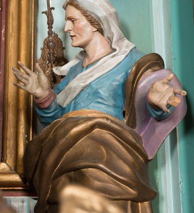 Zdjęcie nr 2: Rzeźba drążona, ustawiona na niskiej podstawie. Święta zwrócona jest w trzech czwartych w prawo, stoi w kontrapoście. Obie ręce ma uniesione, prawą przed piersią, lewą wyciągniętą w bok. Twarz ma szczupłą, pociągłą, o starczych rysach, z wyraźnie podkreśloną linią szczęki, migdałowatymi oczami, osadzonymi blisko siebie, dużym nosie i wąskich, rozchylonych ustach, okoloną jasnobrązowymi włosami, ukrytymi pod welonem. Święta ubrana jest w długą, niebieską tunikę z wąskimi rękawami, o wywiniętym mankiecie, przepasaną w talii oraz złocony płaszcz z jasnofioletową podszewką przerzucony przez lewe ramię, otaczający postać i opadający na lewy bok, na głowie ma biały welon, rozwiany w prawo. Polichromia ciała naturalistyczna.