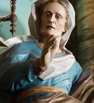 Zdjęcie nr 5: Rzeźba drążona, ustawiona na niskiej podstawie. Święta zwrócona jest w trzech czwartych w prawo, stoi w kontrapoście. Obie ręce ma uniesione, prawą przed piersią, lewą wyciągniętą w bok. Twarz ma szczupłą, pociągłą, o starczych rysach, z wyraźnie podkreśloną linią szczęki, migdałowatymi oczami, osadzonymi blisko siebie, dużym nosie i wąskich, rozchylonych ustach, okoloną jasnobrązowymi włosami, ukrytymi pod welonem. Święta ubrana jest w długą, niebieską tunikę z wąskimi rękawami, o wywiniętym mankiecie, przepasaną w talii oraz złocony płaszcz z jasnofioletową podszewką przerzucony przez lewe ramię, otaczający postać i opadający na lewy bok, na głowie ma biały welon, rozwiany w prawo. Polichromia ciała naturalistyczna.