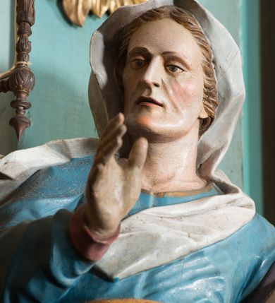 Zdjęcie nr 6: Rzeźba drążona, ustawiona na niskiej podstawie. Święta zwrócona jest w trzech czwartych w prawo, stoi w kontrapoście. Obie ręce ma uniesione, prawą przed piersią, lewą wyciągniętą w bok. Twarz ma szczupłą, pociągłą, o starczych rysach, z wyraźnie podkreśloną linią szczęki, migdałowatymi oczami, osadzonymi blisko siebie, dużym nosie i wąskich, rozchylonych ustach, okoloną jasnobrązowymi włosami, ukrytymi pod welonem. Święta ubrana jest w długą, niebieską tunikę z wąskimi rękawami, o wywiniętym mankiecie, przepasaną w talii oraz złocony płaszcz z jasnofioletową podszewką przerzucony przez lewe ramię, otaczający postać i opadający na lewy bok, na głowie ma biały welon, rozwiany w prawo. Polichromia ciała naturalistyczna.