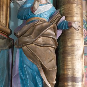 Zdjęcie nr 1: Rzeźba drążona, ustawiona na niskiej podstawie. Święta zwrócona jest w trzech czwartych w prawo, stoi w kontrapoście. Obie ręce ma uniesione, prawą przed piersią, lewą wyciągniętą w bok. Twarz ma szczupłą, pociągłą, o starczych rysach, z wyraźnie podkreśloną linią szczęki, migdałowatymi oczami, osadzonymi blisko siebie, dużym nosie i wąskich, rozchylonych ustach, okoloną jasnobrązowymi włosami, ukrytymi pod welonem. Święta ubrana jest w długą, niebieską tunikę z wąskimi rękawami, o wywiniętym mankiecie, przepasaną w talii oraz złocony płaszcz z jasnofioletową podszewką przerzucony przez lewe ramię, otaczający postać i opadający na lewy bok, na głowie ma biały welon, rozwiany w prawo. Polichromia ciała naturalistyczna.