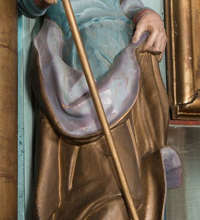 Zdjęcie nr 5: Rzeźba drążona, ustawiona na niskiej podstawie. Święty zwrócony jest w trzech czwartych w lewo, z pochyloną głową, stoi w kontrapoście. Lewą ręką podtrzymuje z przodu płaszcz, w prawej, odchylonej w bok trzyma szuflę pasterską. Twarz ma szczupłą, pociągłą, o starczych rysach, blisko siebie osadzonych oczach skierowanych w dół, dużym nosie, wąskich, otwartych ustach, okoloną długą, falowaną brodą oraz krótkimi, kręconymi włosami. Święty jest ubrany w długą tunikę o wąskich rękawach z wywiniętym brzegiem, przepasaną w talii oraz płaszcz przerzucony przez lewe ramię, opadający na plecy i zawinięty na prawym boku, podtrzymywany lewą dłonią. Polichromia ciała naturalistyczna.