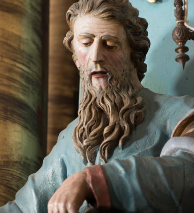 Zdjęcie nr 2: Rzeźba drążona, ustawiona na niskiej podstawie. Święty zwrócony jest w trzech czwartych w lewo, z pochyloną głową, stoi w kontrapoście. Lewą ręką podtrzymuje z przodu płaszcz, w prawej, odchylonej w bok trzyma szuflę pasterską. Twarz ma szczupłą, pociągłą, o starczych rysach, blisko siebie osadzonych oczach skierowanych w dół, dużym nosie, wąskich, otwartych ustach, okoloną długą, falowaną brodą oraz krótkimi, kręconymi włosami. Święty jest ubrany w długą tunikę o wąskich rękawach z wywiniętym brzegiem, przepasaną w talii oraz płaszcz przerzucony przez lewe ramię, opadający na plecy i zawinięty na prawym boku, podtrzymywany lewą dłonią. Polichromia ciała naturalistyczna.