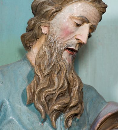 Zdjęcie nr 6: Rzeźba drążona, ustawiona na niskiej podstawie. Święty zwrócony jest w trzech czwartych w lewo, z pochyloną głową, stoi w kontrapoście. Lewą ręką podtrzymuje z przodu płaszcz, w prawej, odchylonej w bok trzyma szuflę pasterską. Twarz ma szczupłą, pociągłą, o starczych rysach, blisko siebie osadzonych oczach skierowanych w dół, dużym nosie, wąskich, otwartych ustach, okoloną długą, falowaną brodą oraz krótkimi, kręconymi włosami. Święty jest ubrany w długą tunikę o wąskich rękawach z wywiniętym brzegiem, przepasaną w talii oraz płaszcz przerzucony przez lewe ramię, opadający na plecy i zawinięty na prawym boku, podtrzymywany lewą dłonią. Polichromia ciała naturalistyczna.