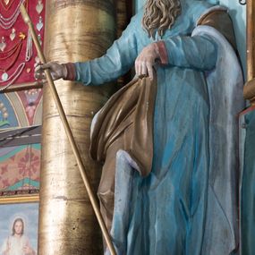 Zdjęcie nr 1: Rzeźba drążona, ustawiona na niskiej podstawie. Święty zwrócony jest w trzech czwartych w lewo, z pochyloną głową, stoi w kontrapoście. Lewą ręką podtrzymuje z przodu płaszcz, w prawej, odchylonej w bok trzyma szuflę pasterską. Twarz ma szczupłą, pociągłą, o starczych rysach, blisko siebie osadzonych oczach skierowanych w dół, dużym nosie, wąskich, otwartych ustach, okoloną długą, falowaną brodą oraz krótkimi, kręconymi włosami. Święty jest ubrany w długą tunikę o wąskich rękawach z wywiniętym brzegiem, przepasaną w talii oraz płaszcz przerzucony przez lewe ramię, opadający na plecy i zawinięty na prawym boku, podtrzymywany lewą dłonią. Polichromia ciała naturalistyczna.