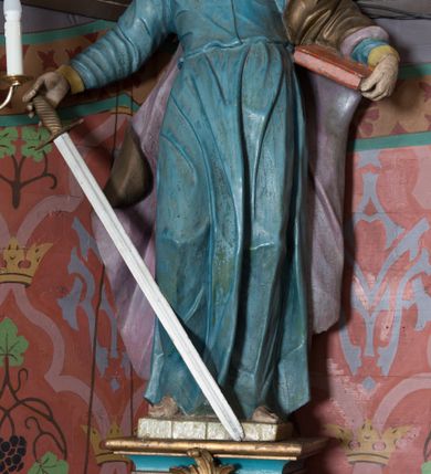 Zdjęcie nr 1: Na konsoli umocowanej do ściany zamykającej prezbiterium, po prawej stronie ołtarza ustawiona figura św. Pawła. Konsola dekorowana rokokowym kartuszem z zasrebrzonym podpisem „ŚW(IĘTY) PAWEŁ”. Rzeźba drążona, zwrócona w trzech czwartych w lewo, w kontrapoście, odchylona w tył. Święty lewą ręką podtrzymuje opartą na boku księgę, w prawej trzyma zwrócony ostrzem w dół miecz. Twarz ma szczupłą, pociągłą, o starczych rysach, blisko siebie osadzonych oczach, garbatym nosie i otwartych, wąskich ustach, okoloną brązową, długą brodą oraz brązowymi, krótkimi i kręconymi włosami. Święty ubrany jest w długą tunikę, o wąskich rękawach, przepasaną w talii oraz złocony płaszcz z jasnofioletową podszewką, przerzucony przez lewe ramie i opadający na plecy, z rozwianą na boki, drapowaną tkaniną. Polichromia ciała naturalistyczna. 