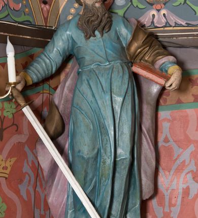 Zdjęcie nr 4: Na konsoli umocowanej do ściany zamykającej prezbiterium, po prawej stronie ołtarza ustawiona figura św. Pawła. Konsola dekorowana rokokowym kartuszem z zasrebrzonym podpisem „ŚW(IĘTY) PAWEŁ”. Rzeźba drążona, zwrócona w trzech czwartych w lewo, w kontrapoście, odchylona w tył. Święty lewą ręką podtrzymuje opartą na boku księgę, w prawej trzyma zwrócony ostrzem w dół miecz. Twarz ma szczupłą, pociągłą, o starczych rysach, blisko siebie osadzonych oczach, garbatym nosie i otwartych, wąskich ustach, okoloną brązową, długą brodą oraz brązowymi, krótkimi i kręconymi włosami. Święty ubrany jest w długą tunikę, o wąskich rękawach, przepasaną w talii oraz złocony płaszcz z jasnofioletową podszewką, przerzucony przez lewe ramie i opadający na plecy, z rozwianą na boki, drapowaną tkaniną. Polichromia ciała naturalistyczna. 