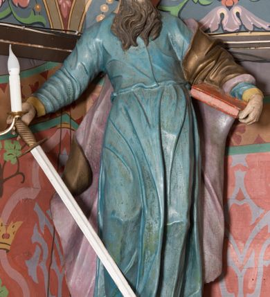 Zdjęcie nr 5: Na konsoli umocowanej do ściany zamykającej prezbiterium, po prawej stronie ołtarza ustawiona figura św. Pawła. Konsola dekorowana rokokowym kartuszem z zasrebrzonym podpisem „ŚW(IĘTY) PAWEŁ”. Rzeźba drążona, zwrócona w trzech czwartych w lewo, w kontrapoście, odchylona w tył. Święty lewą ręką podtrzymuje opartą na boku księgę, w prawej trzyma zwrócony ostrzem w dół miecz. Twarz ma szczupłą, pociągłą, o starczych rysach, blisko siebie osadzonych oczach, garbatym nosie i otwartych, wąskich ustach, okoloną brązową, długą brodą oraz brązowymi, krótkimi i kręconymi włosami. Święty ubrany jest w długą tunikę, o wąskich rękawach, przepasaną w talii oraz złocony płaszcz z jasnofioletową podszewką, przerzucony przez lewe ramie i opadający na plecy, z rozwianą na boki, drapowaną tkaniną. Polichromia ciała naturalistyczna. 