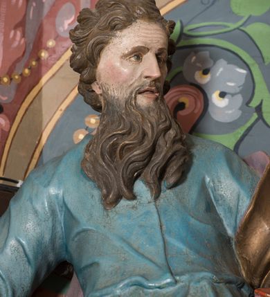 Zdjęcie nr 6: Na konsoli umocowanej do ściany zamykającej prezbiterium, po prawej stronie ołtarza ustawiona figura św. Pawła. Konsola dekorowana rokokowym kartuszem z zasrebrzonym podpisem „ŚW(IĘTY) PAWEŁ”. Rzeźba drążona, zwrócona w trzech czwartych w lewo, w kontrapoście, odchylona w tył. Święty lewą ręką podtrzymuje opartą na boku księgę, w prawej trzyma zwrócony ostrzem w dół miecz. Twarz ma szczupłą, pociągłą, o starczych rysach, blisko siebie osadzonych oczach, garbatym nosie i otwartych, wąskich ustach, okoloną brązową, długą brodą oraz brązowymi, krótkimi i kręconymi włosami. Święty ubrany jest w długą tunikę, o wąskich rękawach, przepasaną w talii oraz złocony płaszcz z jasnofioletową podszewką, przerzucony przez lewe ramie i opadający na plecy, z rozwianą na boki, drapowaną tkaniną. Polichromia ciała naturalistyczna. 