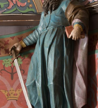 Zdjęcie nr 3: Na konsoli umocowanej do ściany zamykającej prezbiterium, po prawej stronie ołtarza ustawiona figura św. Pawła. Konsola dekorowana rokokowym kartuszem z zasrebrzonym podpisem „ŚW(IĘTY) PAWEŁ”. Rzeźba drążona, zwrócona w trzech czwartych w lewo, w kontrapoście, odchylona w tył. Święty lewą ręką podtrzymuje opartą na boku księgę, w prawej trzyma zwrócony ostrzem w dół miecz. Twarz ma szczupłą, pociągłą, o starczych rysach, blisko siebie osadzonych oczach, garbatym nosie i otwartych, wąskich ustach, okoloną brązową, długą brodą oraz brązowymi, krótkimi i kręconymi włosami. Święty ubrany jest w długą tunikę, o wąskich rękawach, przepasaną w talii oraz złocony płaszcz z jasnofioletową podszewką, przerzucony przez lewe ramie i opadający na plecy, z rozwianą na boki, drapowaną tkaniną. Polichromia ciała naturalistyczna. 