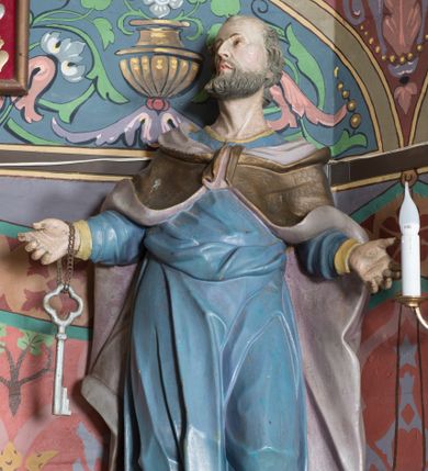 Zdjęcie nr 1: Na konsoli umocowanej do ściany zamykającej prezbiterium, po lewej stronie ołtarza ustawiona jest figura św. Piotra. Konsola dekorowana rokokowym kartuszem z zasrebrzonym napisem „ŚW(IĘTY) PIOTR”. Rzeźba jest drążona, zwrócona w trzech czwartych w prawo, w kontrapoście, z obiema rękami rozłożonymi na boki. Na prawym nadgarstku ma zawieszony klucz. Święty ma szczupłą i pociągłą twarz z wysokim czołem, osadzonymi blisko siebie oczami, lekko garbatym nosem i otwartymi, wąskimi ustami, okoloną krótką, kręconą, wysuniętą do przodu brodą oraz kręconymi włosami i puklem nad czołem, na głowie łysina. Piotr ubrany jest w długą, niebieską tunikę, o wąskich rękawach, przepasaną w talii oraz narzucony na ramiona i zapięty na piersi złocony płaszcz z jasnofioletową podszewką. Polichromia ciała naturalistyczna.