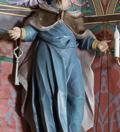 Zdjęcie nr 2: Na konsoli umocowanej do ściany zamykającej prezbiterium, po lewej stronie ołtarza ustawiona jest figura św. Piotra. Konsola dekorowana rokokowym kartuszem z zasrebrzonym napisem „ŚW(IĘTY) PIOTR”. Rzeźba jest drążona, zwrócona w trzech czwartych w prawo, w kontrapoście, z obiema rękami rozłożonymi na boki. Na prawym nadgarstku ma zawieszony klucz. Święty ma szczupłą i pociągłą twarz z wysokim czołem, osadzonymi blisko siebie oczami, lekko garbatym nosem i otwartymi, wąskimi ustami, okoloną krótką, kręconą, wysuniętą do przodu brodą oraz kręconymi włosami i puklem nad czołem, na głowie łysina. Piotr ubrany jest w długą, niebieską tunikę, o wąskich rękawach, przepasaną w talii oraz narzucony na ramiona i zapięty na piersi złocony płaszcz z jasnofioletową podszewką. Polichromia ciała naturalistyczna.